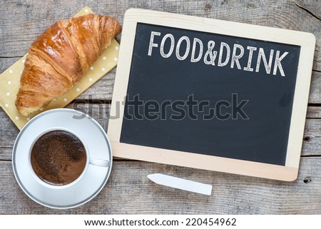 Overhead view of a fresh cup of coffee and a flaky croissant on an old school slate over a rustic wooden background.With text Food and Drink on blackboard