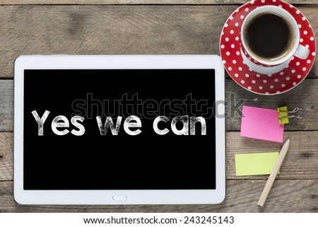 Yes we can phrase. Tablet computer with stickers,cup of coffee and yes we can phrase on the wooden table