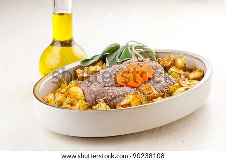 Roasted  meet and potatoes with sage and carrots garnish in dish over a white wood table and an oil bottle on side