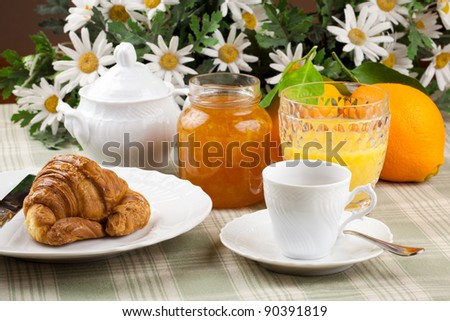 breakfast including coffee cup, pastry, croissant, orange marmalade and orange juice over a country table cloth with bunch of daisies and fresh oranges in background