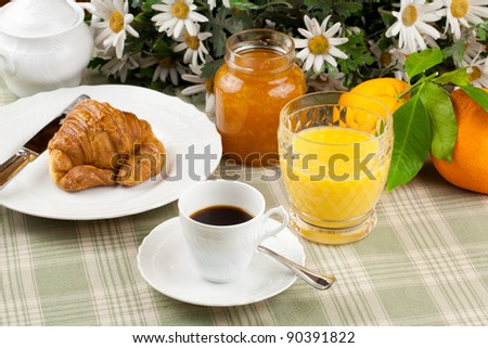 breakfast including coffee cup, pastry, croissant, orange marmalade and orange juice served over a country table cloth with bunch of daisies and fresh oranges in background