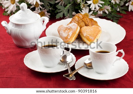Two coffee cup and two heart shaped pastry over a red table cloth with daisies bouquet in background