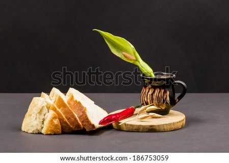 Still life composition with wooden kitchen cutting board, bread, red pepper and ceramic pot with Arum flower on dark background