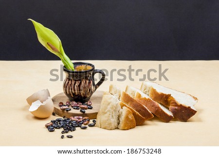 Still life composition with wooden kitchen cutting board, beans, egg shell, bread and ceramic pot with Arum flower on dark background. Image has grain texture visible on its maximum size