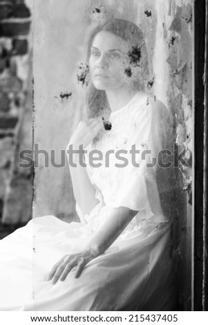 Beautiful girl shot through a dirty glass in an abandoned house wearing an old fashioned wedding dress.  Photo has grain texture visible on its maximum size.  Artistic black and white photography