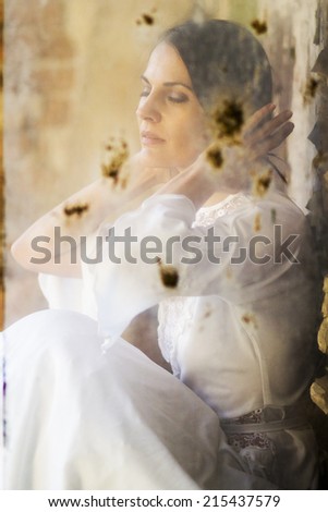 Beautiful girl shot through a dirty glass in an abandoned house wearing an old fashioned wedding dress.  Photo has grain texture visible on its maximum size.  Artistic  photography