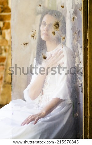 Beautiful girl shot through a dirty glass in an abandoned house wearing an old fashioned wedding dress.  Photo has grain texture visible on its maximum size.  Artistic photography