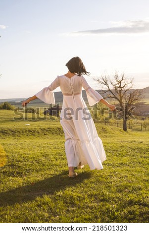 Brunette girl posing in the field with tree, wearing a white wedding dress posing with sunset light. Photo has grain texture visible on its maximum size. Artistic photography