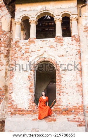 Beautiful, brunette woman in an old, abandoned house, wearing an orange dress, looking sad and melancholic. Photo has grain texture visible on its maximum size. Artistic photography