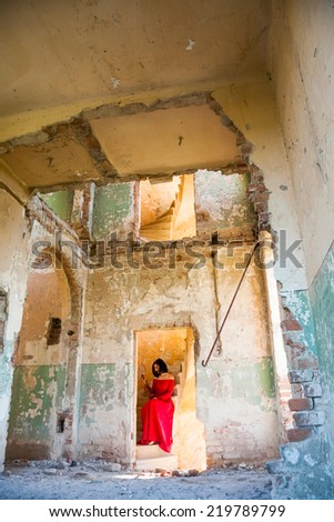 Beautiful, brunette woman in an old, abandoned house, wearing a red dress, looking sad and melancholic. Photo has grain texture visible on its maximum size. Artistic photography