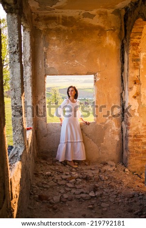 Beautiful, brunette woman in an old, abandoned house, wearing a wedding dress, posing with sunset light. Photo has grain texture visible on its maximum size. Artistic photography