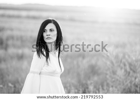 Brunette girl in the field, wearing a white dress, looking sad and worried. Photo has grain texture visible on its maximum size. Artistic black and white photography