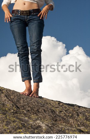 Woman legs, feet and hands with jeans, on a rock, with natural background and fluffy, white clouds