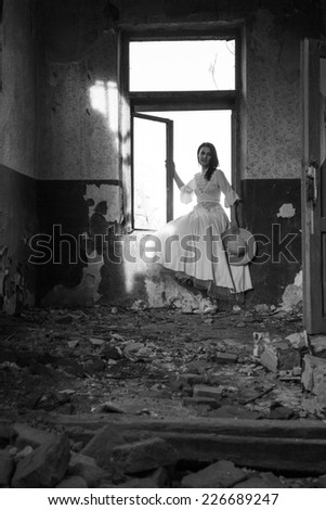 Sad mood in an old, abandoned house with girl wearing an old fashioned wedding dress with natural light. Photo has grain texture visible on its maximum size. Artistic, black and white photography