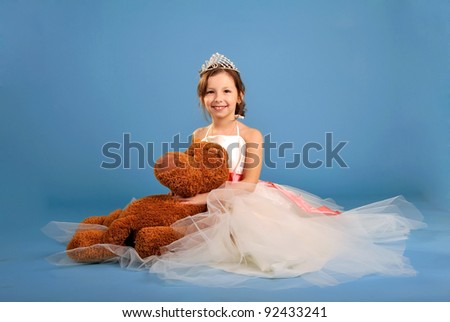 Little beautiful girl sitting in white spruce dress and crown with toy bear in hands