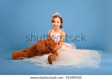 Little beautiful girl sitting in white spruce dress and crown with toy bear in hands