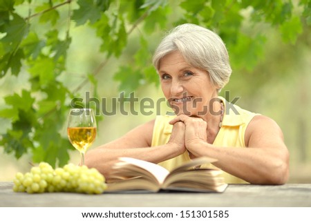 Old woman sitting at a table, drinking wine and reading a book