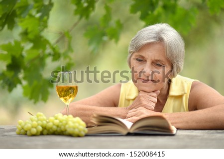 Old woman sitting at a table, drinking wine and reading a book