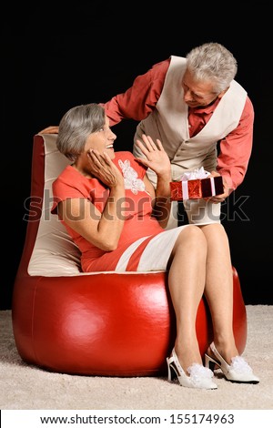 Happy senior man giving gift to his wife who sitting on red armchair in dark studio