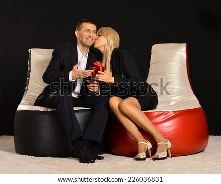 Man and woman sitting on black and red armchairs with gift in studio