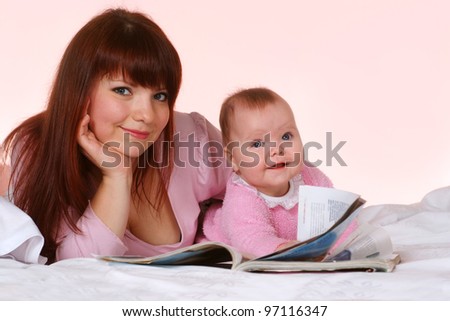 Beautiful charming mother and daughter lying in bed on a pink background with a magazine