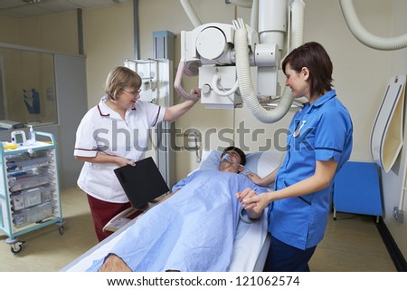 radiographer placing X Ray plate under teenage male patient lying under X Ray machine while nurse reassures him