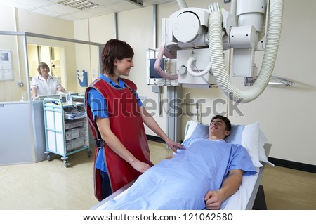 young nurse wearing lead apron smiling reassuring teenage patient on X Ray table with radiographer behind glass screen watching and controlling X ray machine