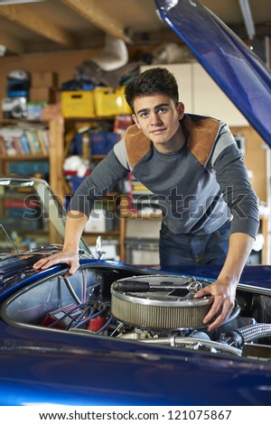 teenage boy leaning over the engine of a classic car in garage looking at camera