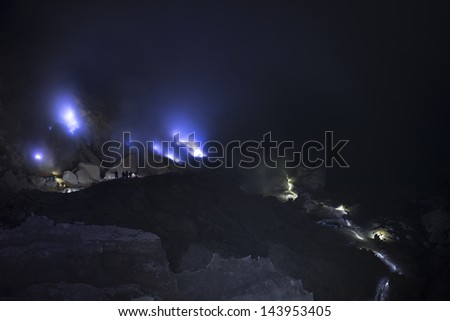 A sulfur miner stands inside the crater of the Kawah Ijen volcano at night.  a flow of liquid sulfur which has caught fire and burns with an blue flame.