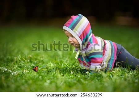 Curious baby in striped dress crawling on a green lawn to the butterfly. The child first saw a butterfly and looks very surprised