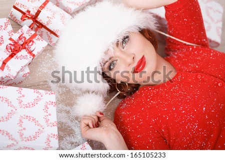 Beautiful young woman in a red sweater and white fur hat lying on the floor with many gift boxes. Christmas portrait.