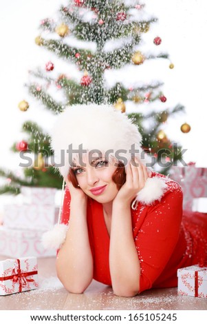 Beautiful young woman in a red sweater and white fur hat lying on the floor near Christmas tree