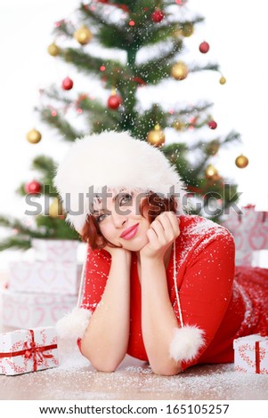 Beautiful young woman in a red sweater and white fur hat lying on the floor near Christmas tree