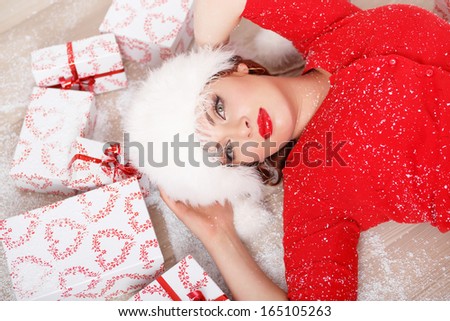 Beautiful young woman in a red sweater and white fur hat lying on the floor with many gift boxes. Christmas portrait.