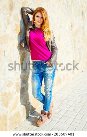 Stylish young girl with red hair posing in jeans and leather jacket against rock wall, street fashion photo