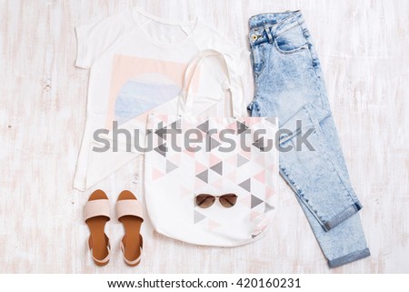White t-shirt with print, light blue ripped boyfriend jeans, beige sandals, sunglasses, cotton textile bag on white wooden background. Overhead view of woman\'s casual outfits. Flat lay, top view.