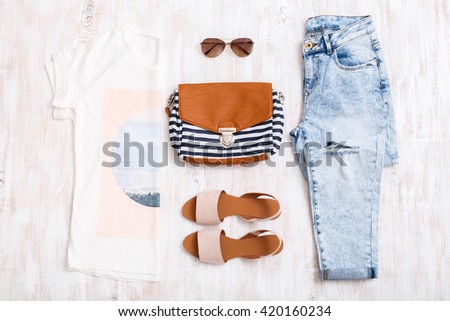 White t-shirt with print, light blue ripped boyfriend jeans, beige sandals, sunglasses, textile striped bag on white wooden background. Overhead view of woman\'s casual outfits. Flat lay, top view.