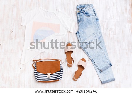 White t-shirt with print, light blue ripped boyfriend jeans, beige sandals, textile striped bag on white wooden background. Overhead view of woman\'s casual outfits. Flat lay, top view.