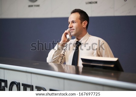 Reception of hotel, desk clerk, man taking a call and smiling .Night light.