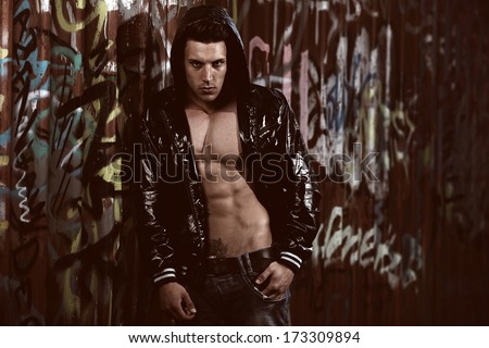 Young handsome macho man with open jacket revealing muscular chest and abs in industrial garage with graffiti