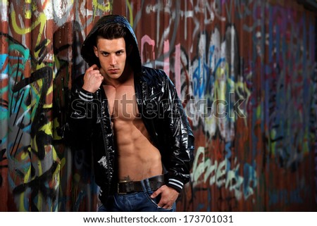 Young handsome macho man with open jacket revealing muscular chest and abs in industrial garage with graffiti .Fashion colors.