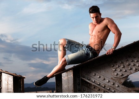 Handsome young muscle man shirtless with hand on rusty metal structure, looking in camera .Fashion colors