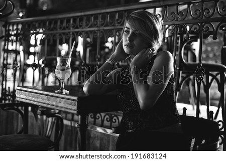 Attractive blonde woman in elegant black dress sitting on bar stool. Gorgeous blonde model posing provocatively in vintage bar .B&W
