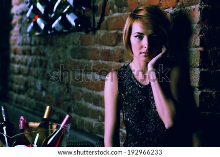 Attractive blonde woman in elegant black dress sitting on bar stool. Gorgeous blonde model posing provocatively in vintage bar .Fashion colors.