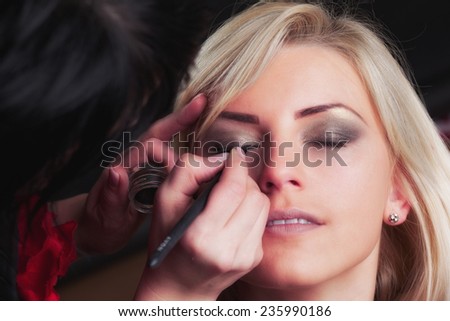 Make-up artist applying bright base color eyeshadow on model\'s eye and holding a shell with eyeshadow on background, close up