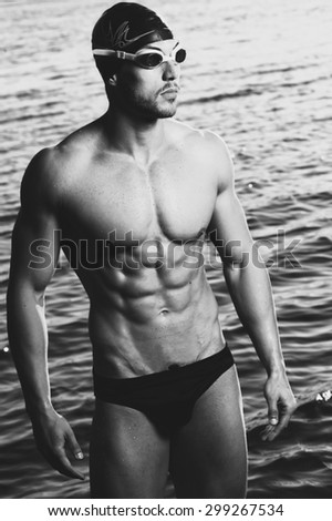 Muscled swimmer man with cap and glasses outdoor at a lake with blue cloudy sky.  Extreme fitness sport. Fashion colors.