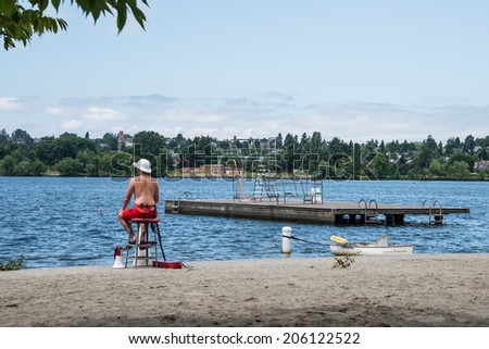 SEATTLE, WA - JULY 2, 2014:  Water Safety and Recreation: Lifeguard on duty on beach overlooking lake with swimming area and diving platform, Green Lake Park, Seattle Washington.