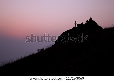 dramatic silhouette of a young male hiker standing, one climbing and one sitting on a big rock near the top of a hill in early morning with purple pink cloudy sky as background