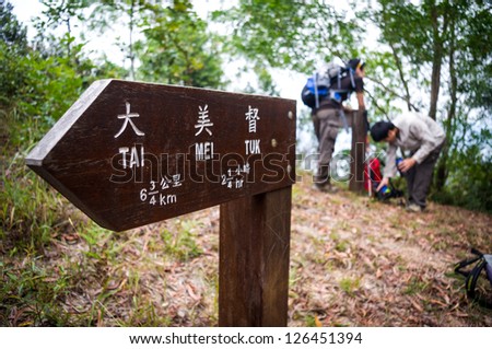 a wooden direction sign pointing left with chinese and english name of tai mei tuk, in Hong Kong, and details of distance and time needed.  2 unrecognizable blurred young male hikers at back resting