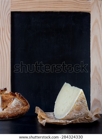 Soft cheese wedge and piece of rustic bread loaf in front of blank old blackboard carte.